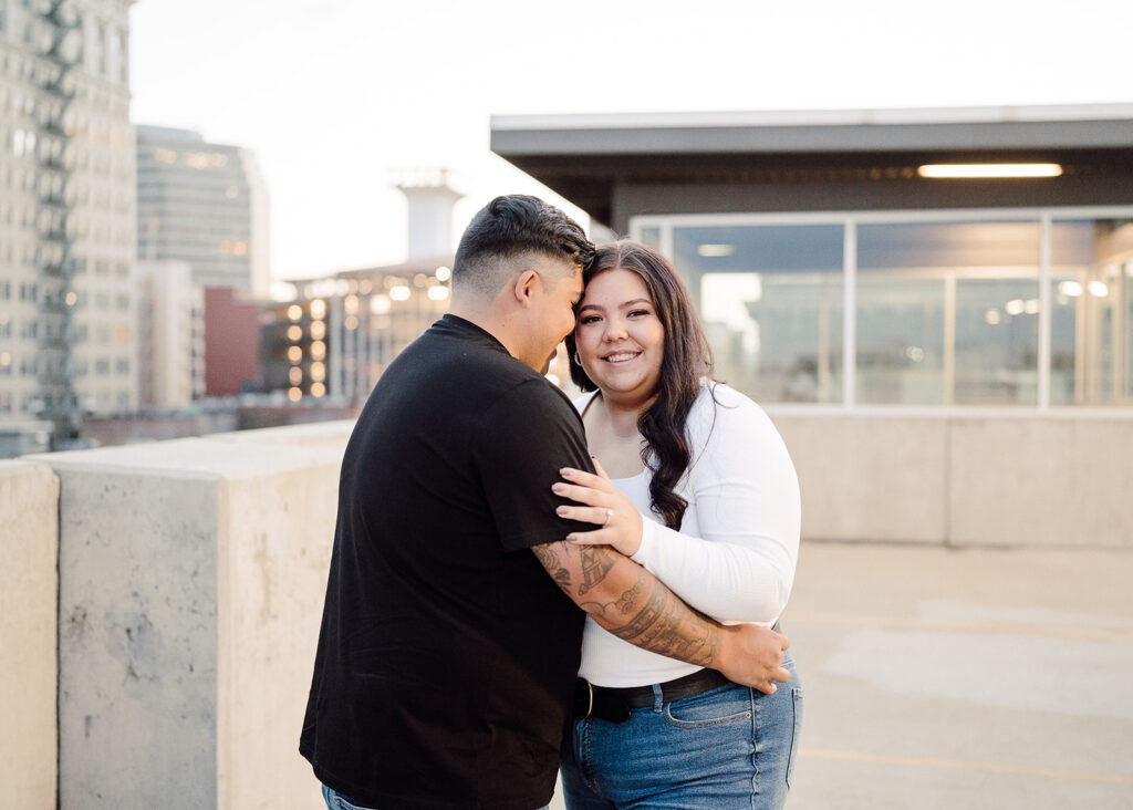 An engaged couple smiling and holding hands while walking down a rooftop in downtown Spokane. The urban setting features tall buildings, streetlights, and colorful storefronts, adding a vibrant and lively backdrop to the couple's romantic moments captured in the photos
